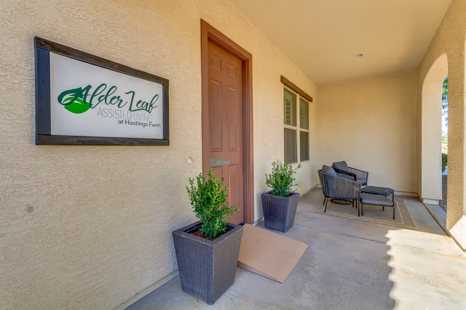 Covered entrance porch of Alder Leaf assisted living with a wall sign, potted plants, and outdoor seating.