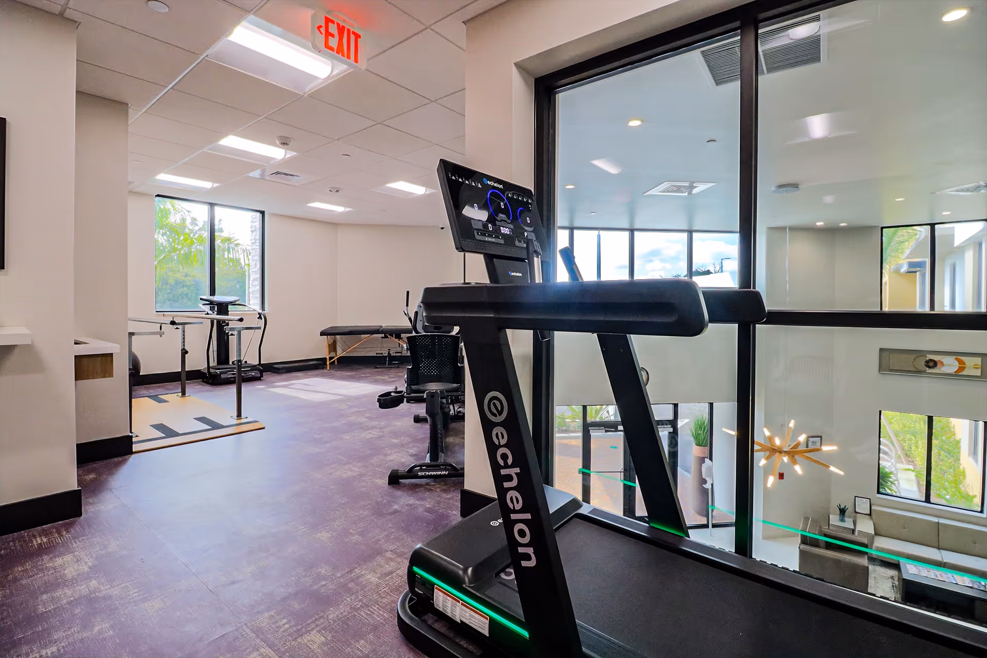 A fitness room with exercise equipment including a treadmill, stationary bike, and parallel bars. The room has large windows letting in natural light and a view into a lower level with modern decor and lighting fixtures.