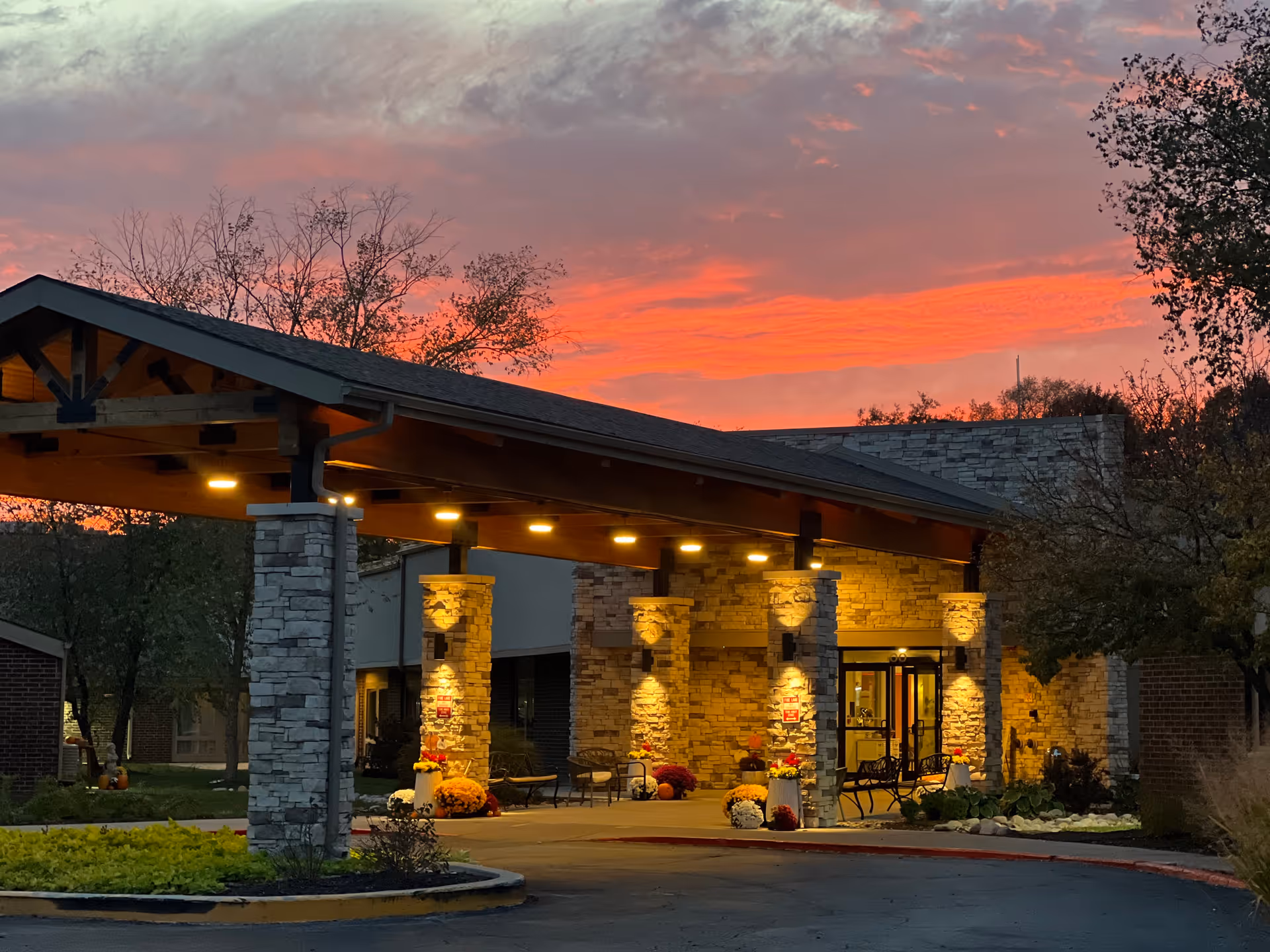 The entrance of The Grand Royale Senior Living facility at sunset, featuring a covered driveway with stone pillars illuminated by warm lights, surrounded by trees and landscaping.