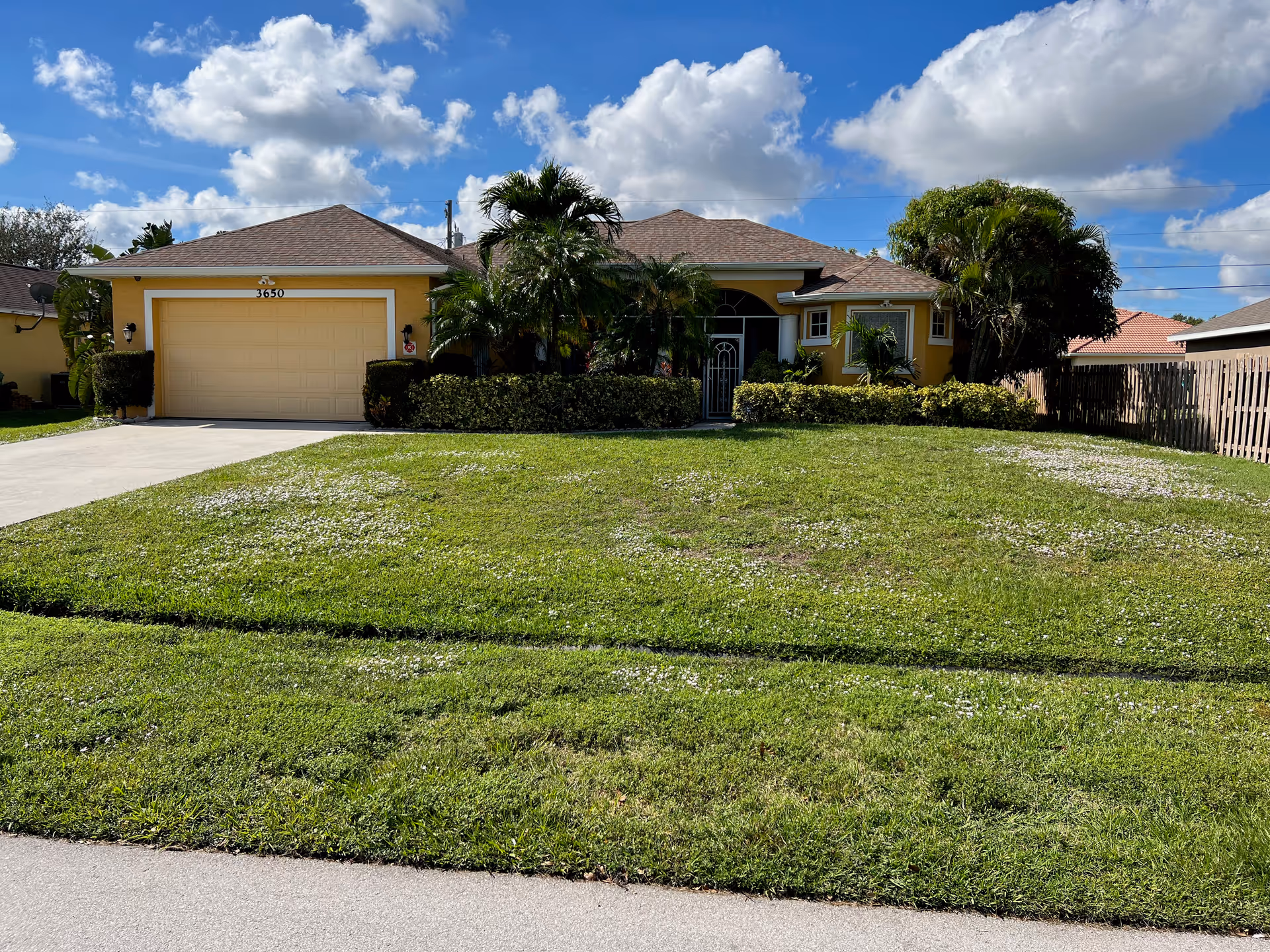 Single-story yellow house with a two-car garage, manicured lawn, and palm trees under a blue sky.