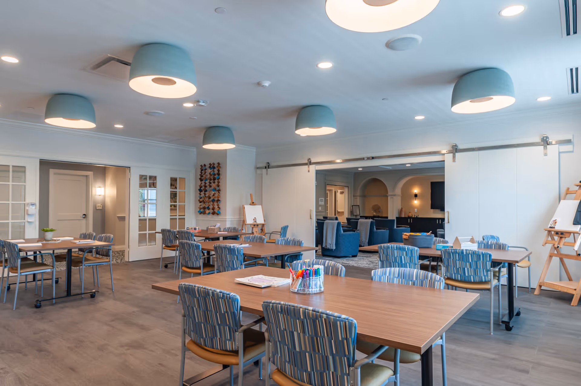 A spacious, well-lit activity room in Arbor Terrace Marlton featuring multiple tables with chairs arranged around them. The room has modern ceiling lights, wooden floors, and a neutral color palette. In the background, there is a lounge area with blue armchairs and a TV mounted on the wall. Art supplies and an easel are visible, suggesting a creative or recreational space.