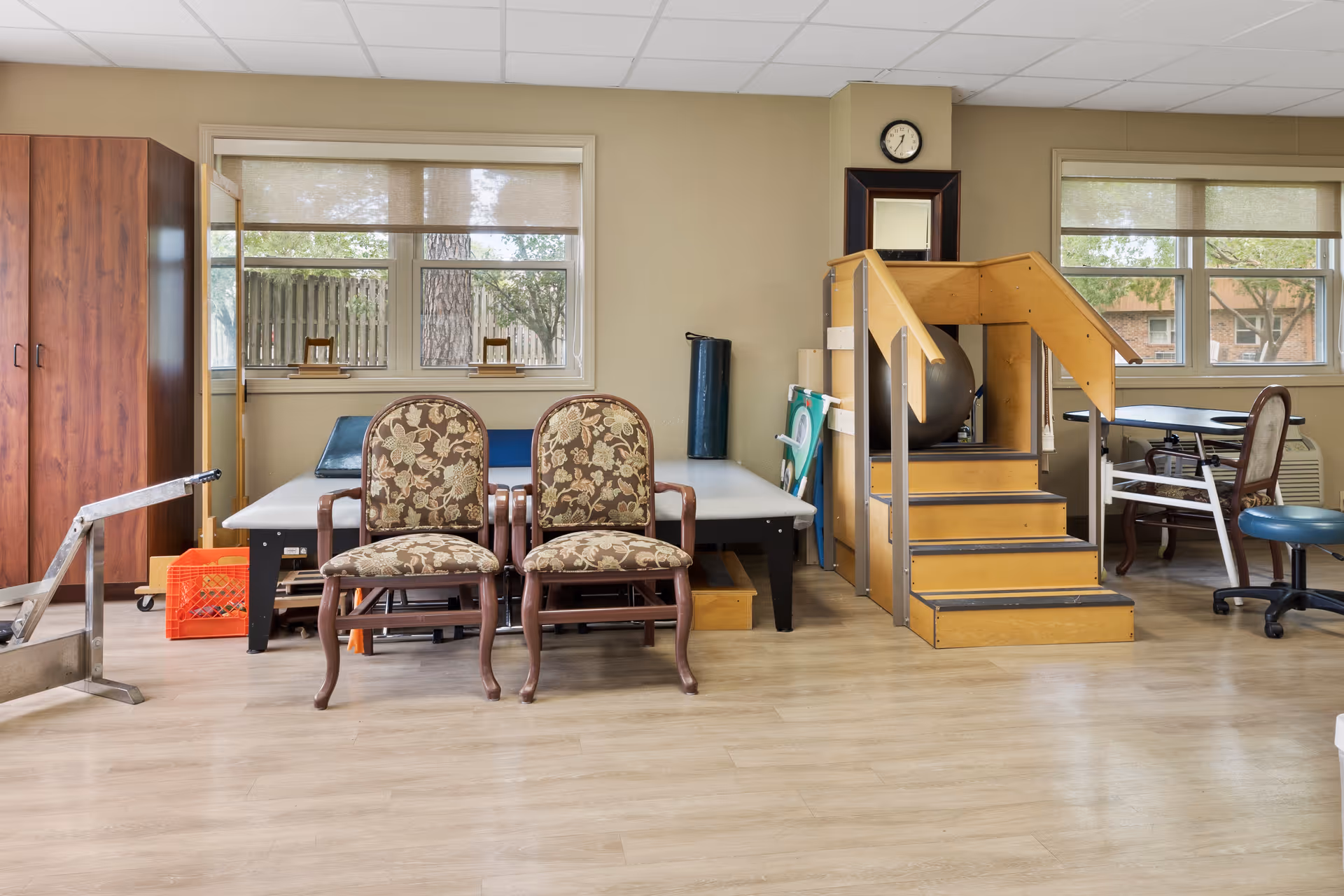 A physical therapy or rehabilitation room with two floral upholstered chairs in front of a padded therapy table, a small wooden staircase with handrails, exercise equipment including a large exercise ball, and windows letting in natural light.