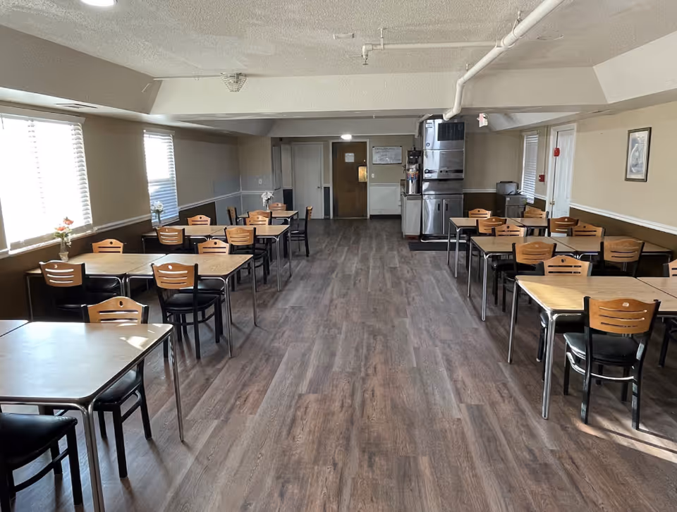A dining room with multiple wooden tables and chairs arranged neatly on a wood-patterned floor. The room has beige walls, several windows with blinds letting in natural light, and a beverage station with coffee machines and supplies at the far end.