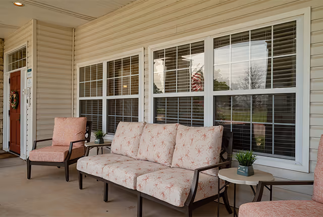 Covered outdoor patio area with cushioned seating including a sofa and two armchairs, small side tables with potted plants, and large windows with white blinds behind the seating.