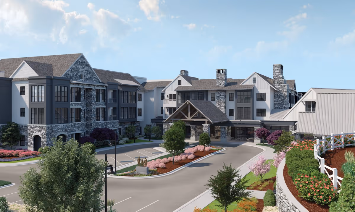 Front exterior of a multi-story senior living facility with stone accents, a covered porte-cochere entrance, landscaped driveway and flowering shrubs.