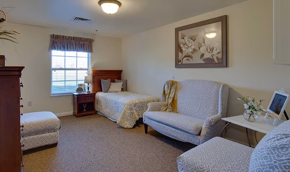 A cozy senior living bedroom with a single bed covered in a patterned quilt, a wooden nightstand with a lamp and clock, a cushioned loveseat with a throw blanket, a matching cushioned chair and ottoman, a small side table with a vase of flowers and a framed photo, and a window with blinds and a valance.