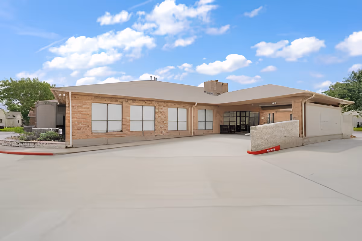 Exterior view of a single-story building with a covered entrance and multiple windows, surrounded by a paved driveway and some greenery under a partly cloudy sky.