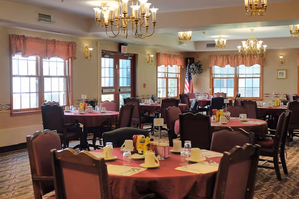 A dining room with multiple round tables covered with red tablecloths, each set with white cups, glasses, and condiments. The room has large windows with red valances, chandeliers hanging from the ceiling, and an American flag in the corner near the windows.
