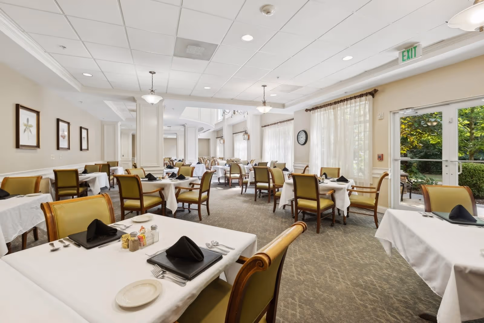 A bright and spacious dining room with multiple tables covered in white tablecloths, each set with black napkins, plates, and silverware. The room features beige walls, framed artwork, large windows with sheer curtains, and an exit door leading outside to a garden area. Comfortable wooden chairs with green cushions surround the tables.