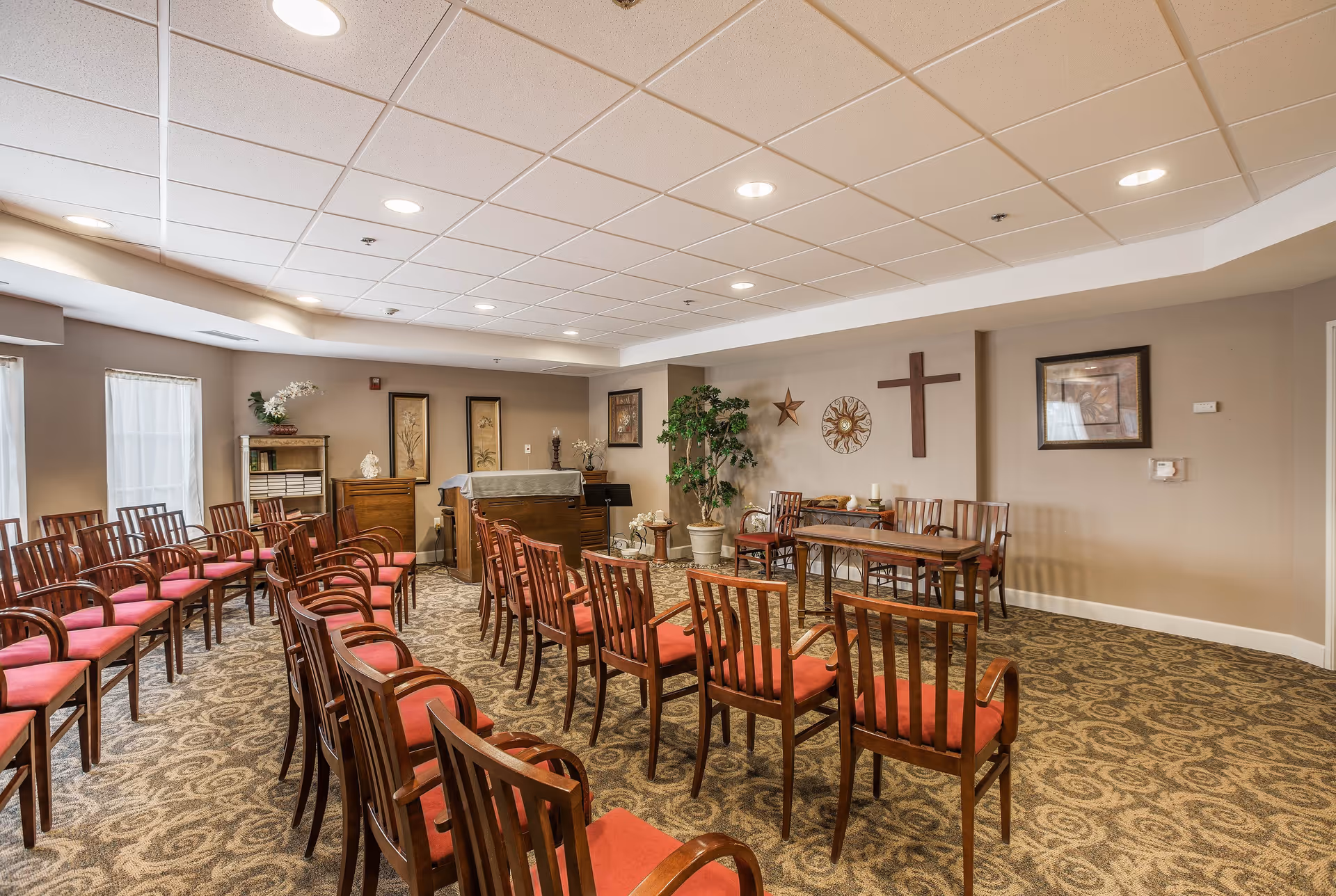 Interior view of a chapel or meeting room with rows of wooden chairs with red cushions arranged facing a small altar area. The room has beige walls decorated with framed pictures, a cross, a star, and a circular wall ornament. There is a carpeted floor with a patterned design and a ceiling with recessed lighting.