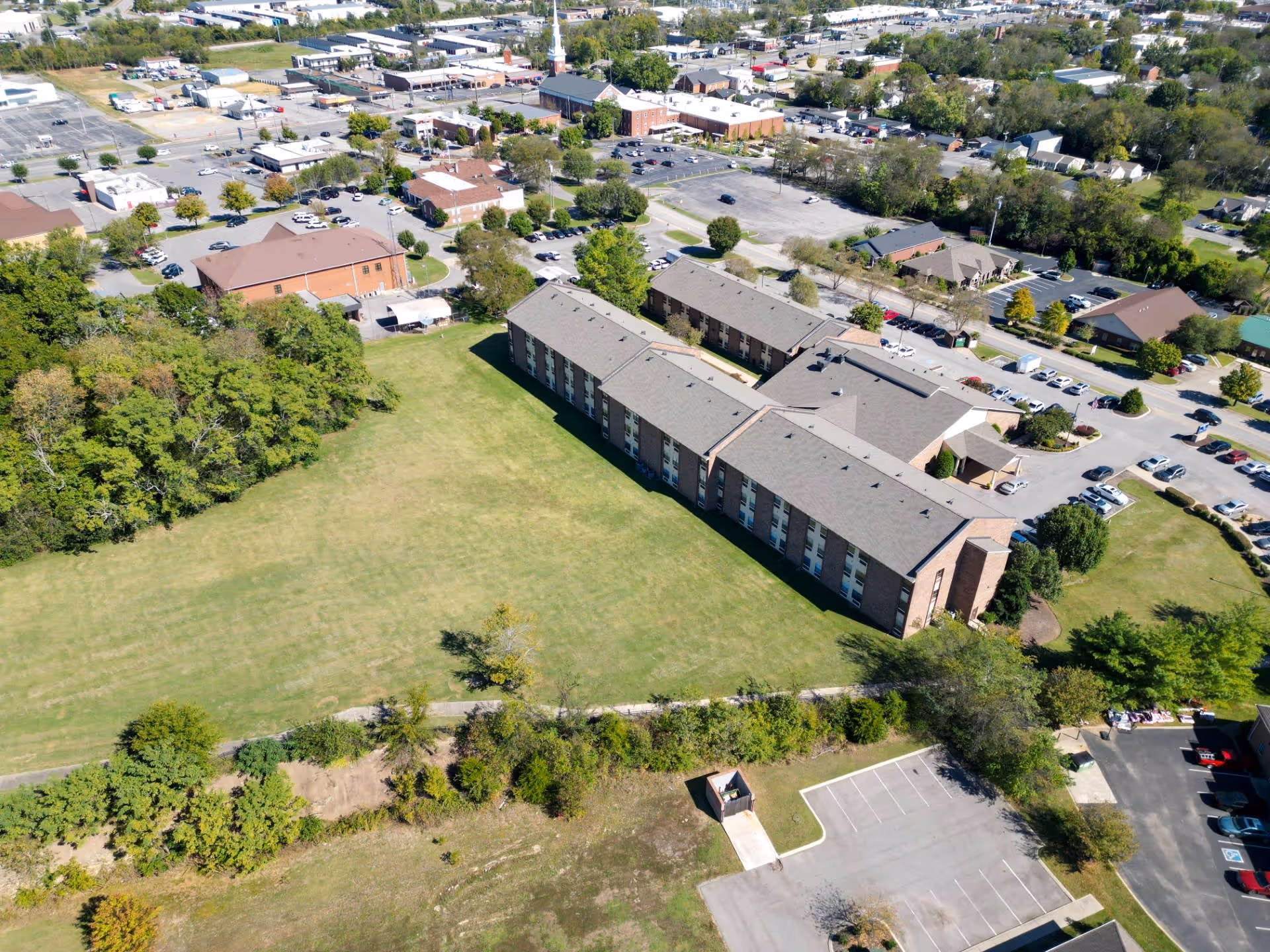 Aerial view of Park Place Retirement Community showing a large multi-story building surrounded by green lawns, trees, parking lots, and nearby roads with other buildings in the background.