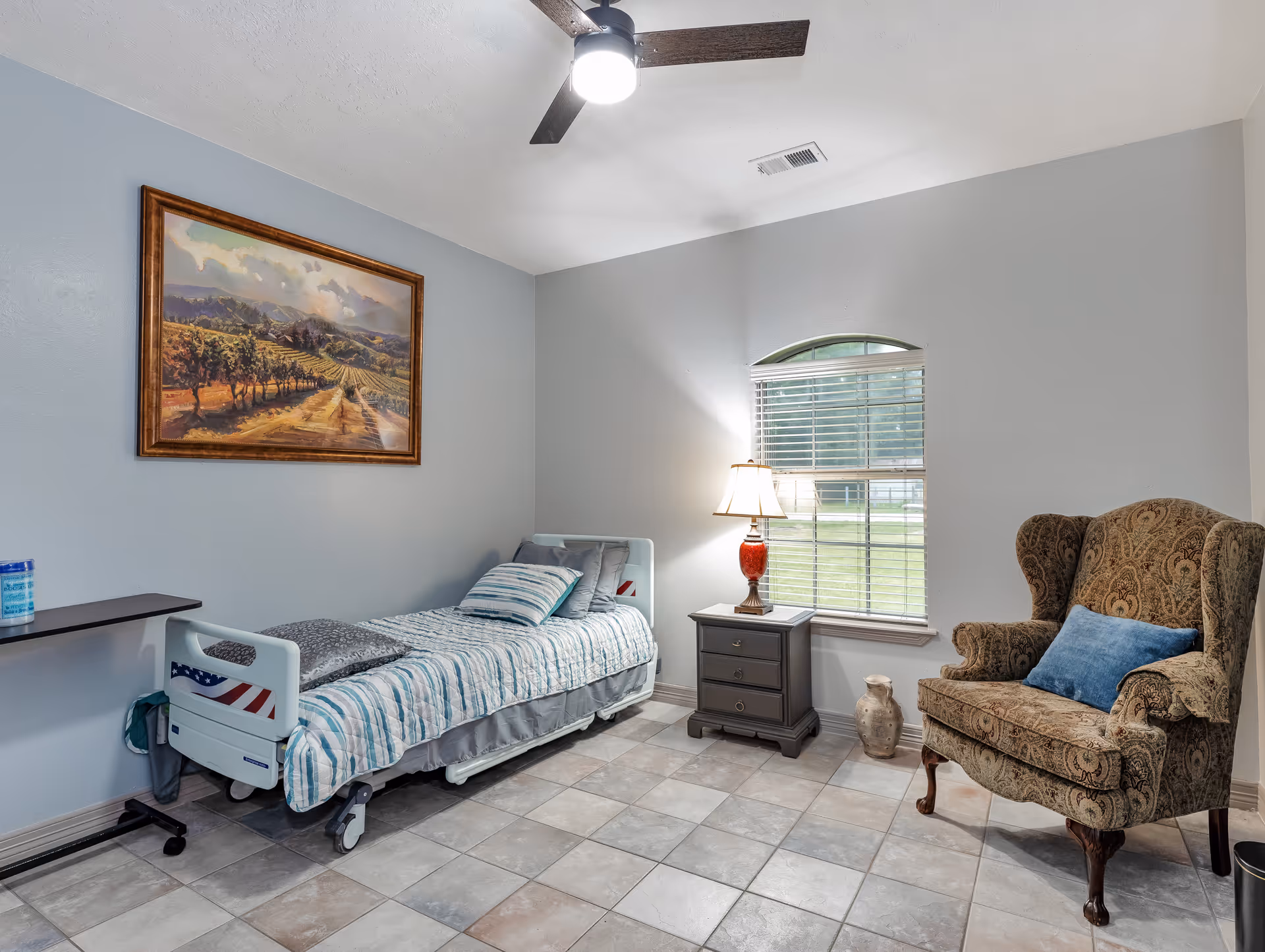 A small, neatly arranged bedroom with a single hospital-style bed covered in striped bedding, a bedside table with a lamp, a patterned armchair with a blue cushion, a window with blinds, and a framed landscape painting on the wall.