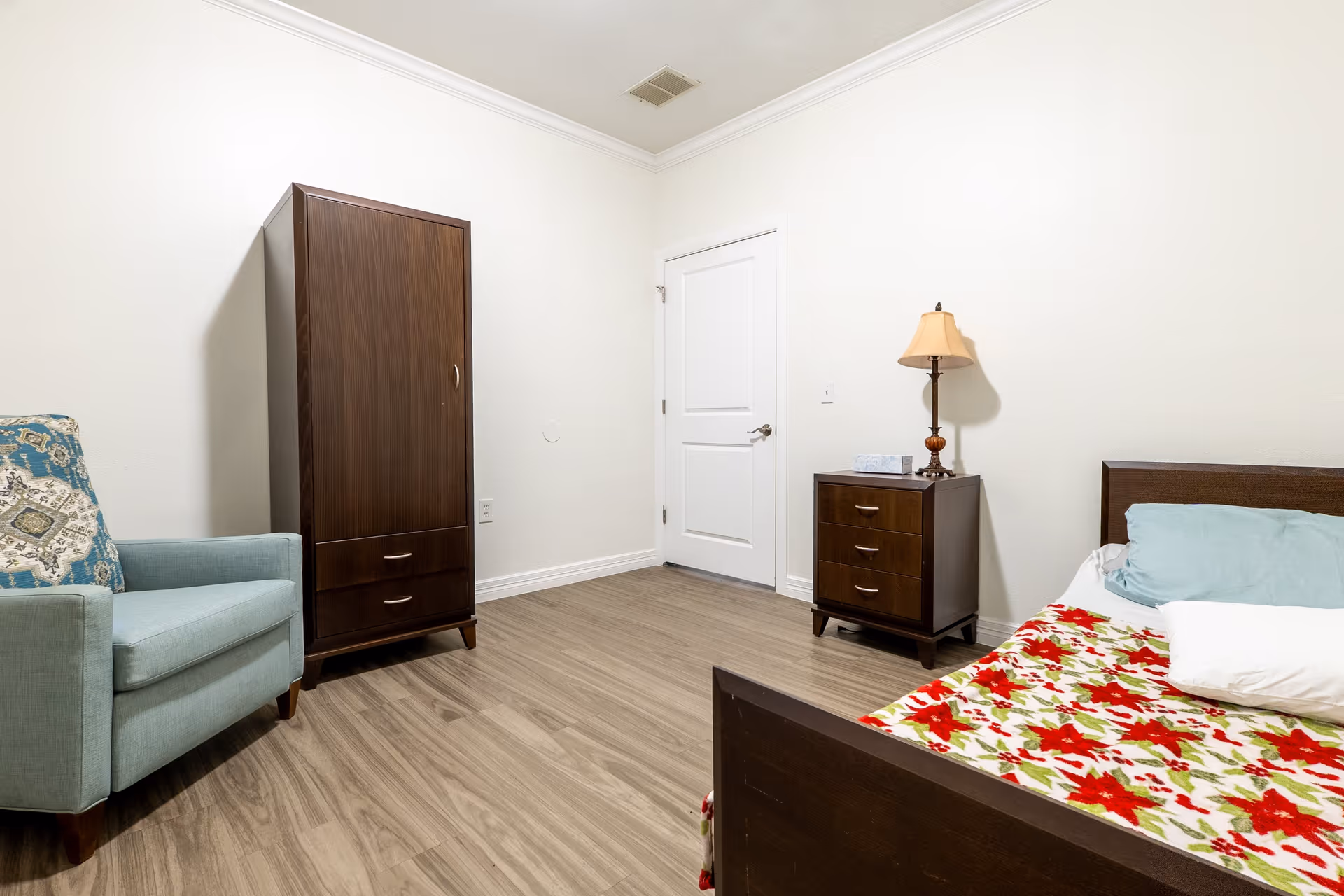 A simple bedroom with light wood flooring, white walls, and white ceiling. The room contains a dark wood bed with a colorful red and green floral blanket, a light blue armchair with a patterned cushion, a dark wood wardrobe, and a matching dark wood nightstand with a lamp and a box of tissues. A closed white door is visible on the far wall.