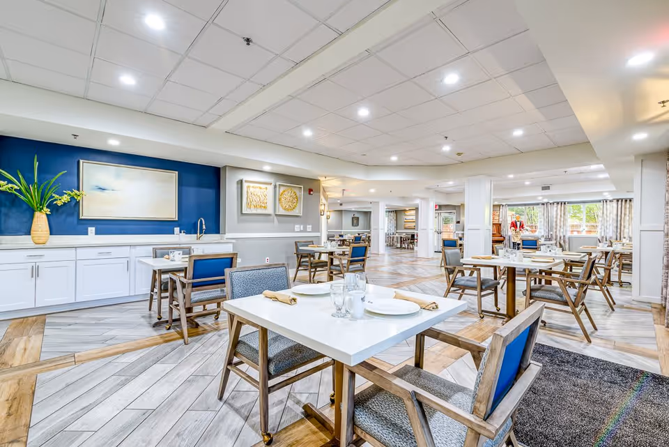 Bright and spacious dining room in a senior living facility with multiple tables set with plates, glasses, and napkins. The room features modern wooden chairs with blue cushions, light wood and tile flooring, white cabinetry along one wall with decorative artwork and a vase with green plants, and large windows allowing natural light to enter.
