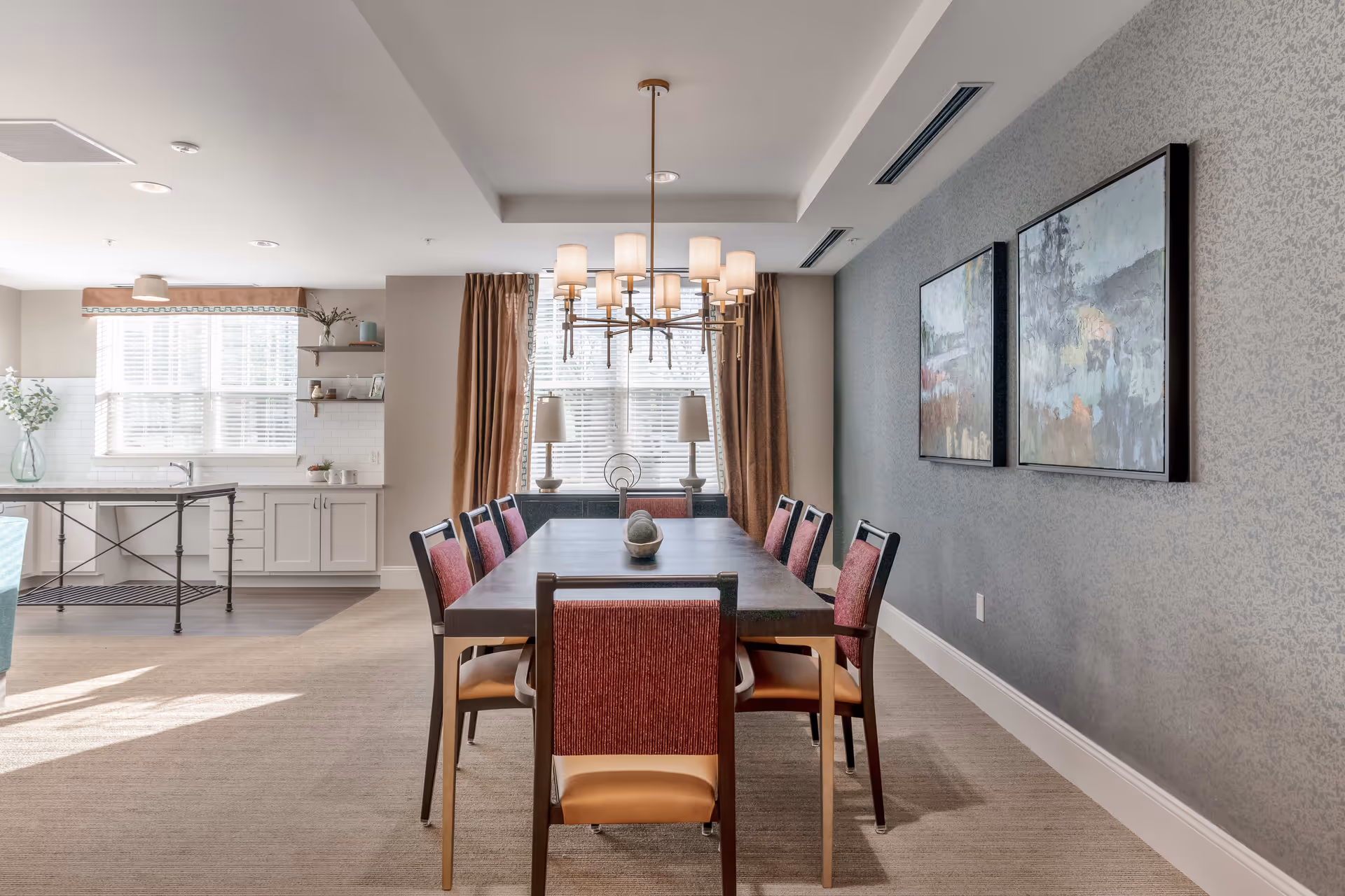 A bright dining room with a long rectangular table surrounded by eight chairs with red backs and tan seats. A modern chandelier with multiple lights hangs above the table. The room has large windows with brown curtains and white blinds, two table lamps on a sideboard, and two abstract paintings on a patterned gray wall. The adjacent kitchen area features white cabinets, a large window, and a kitchen island with a metal frame.