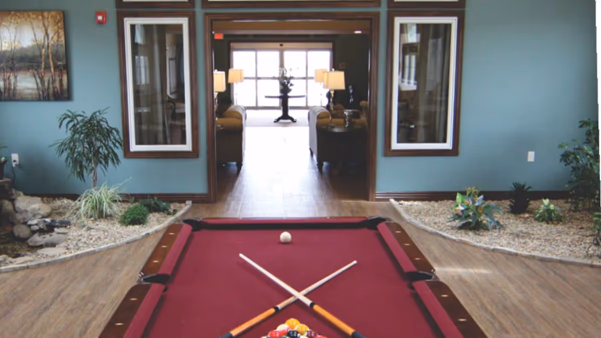 Indoor common area featuring a red pool table in the foreground with potted plants and a seating/lounge area visible through an open doorway.