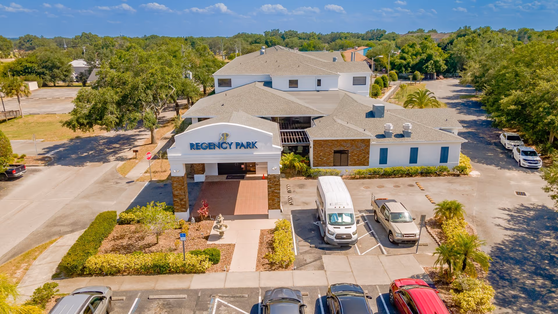 Aerial view of Regency Park Assisted Living & Memory Care building with a parking lot in front, surrounded by trees and greenery under a blue sky.