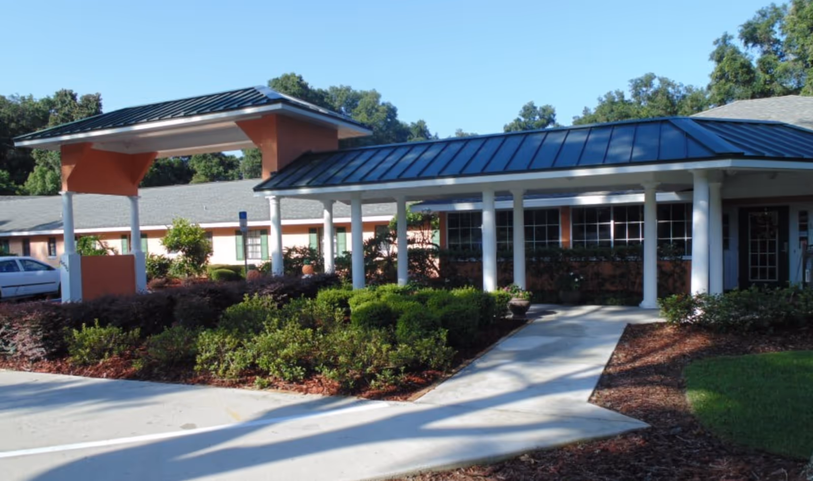 Exterior view of Hampton Manor Assisted Living at Belleview showing a covered entrance with white pillars, a walkway, landscaped bushes, and a building with green window shutters under a clear blue sky.
