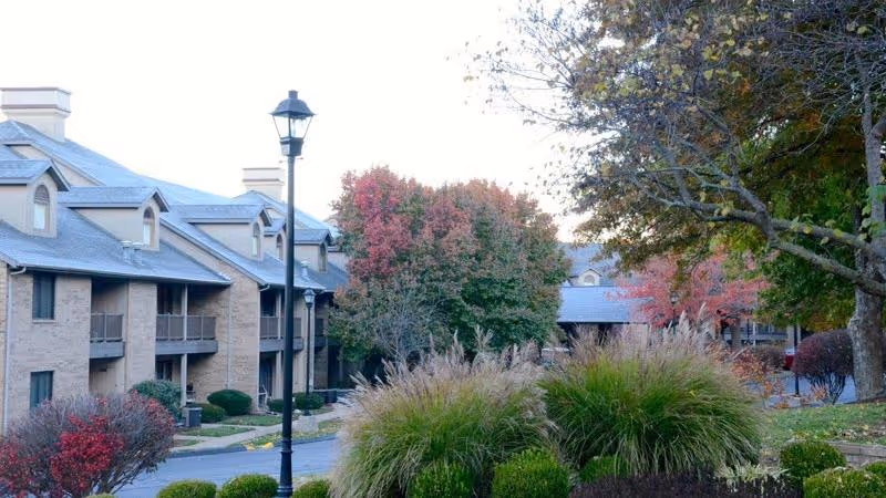 Front exterior view of a multi-story brick senior living building with balconies, lamp posts, and landscaped trees and shrubs in autumn.