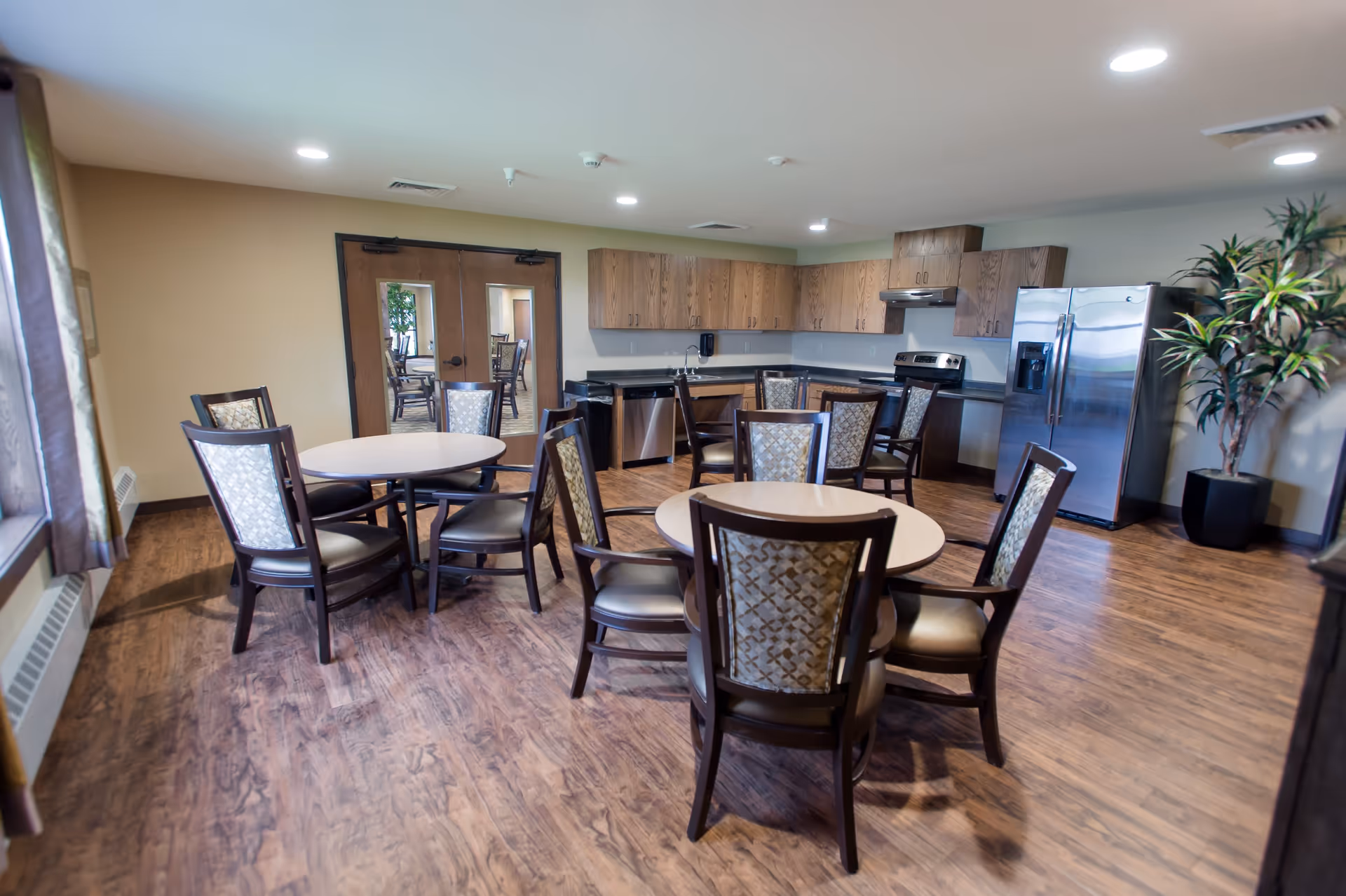 A bright and clean communal dining area with round tables and cushioned chairs. The room features wood flooring, a kitchen area with wooden cabinets, a stainless steel refrigerator, stove, and dishwasher. There are double wooden doors with glass panels and a large potted plant in the corner.