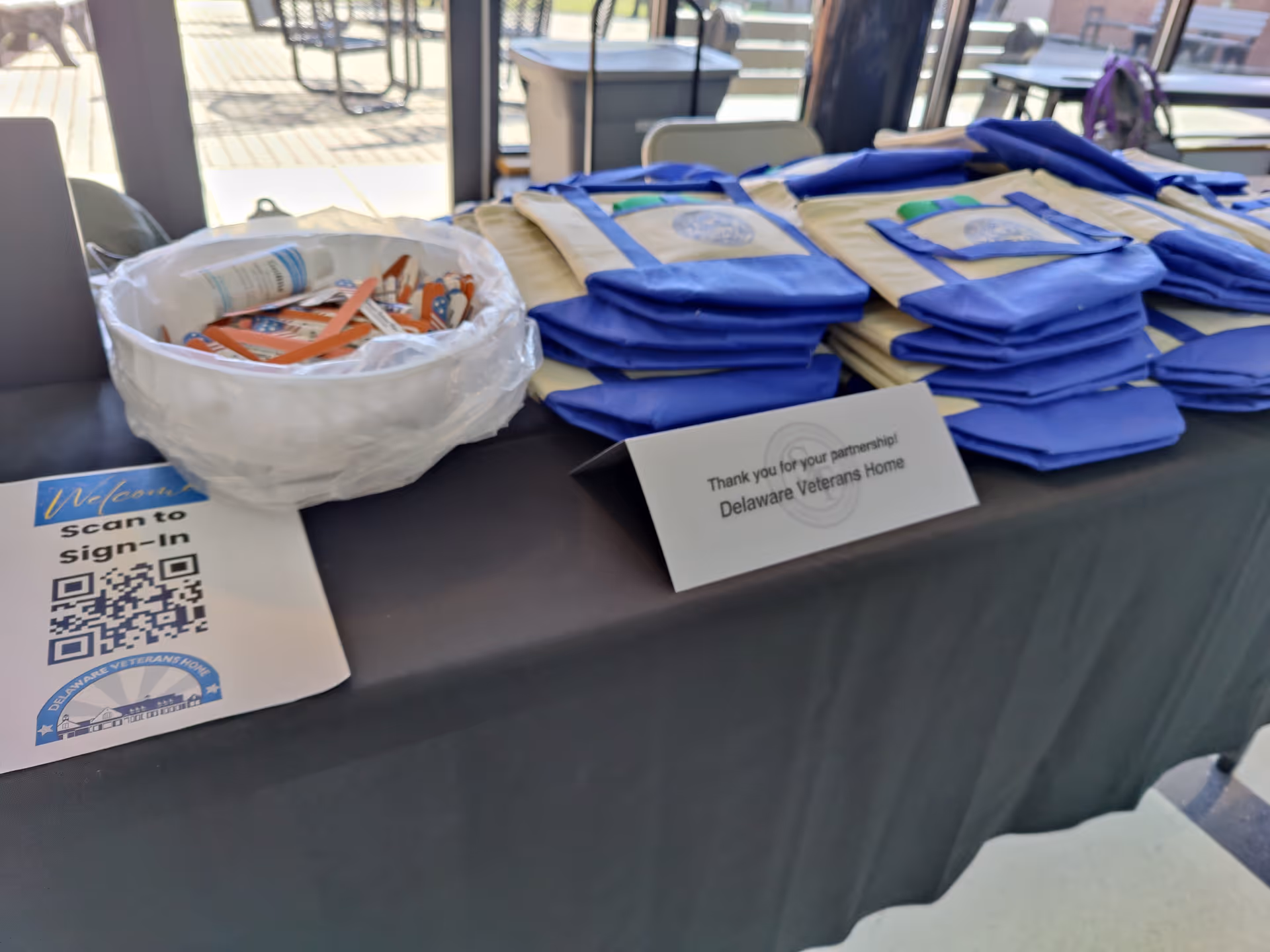 Indoor table displaying stacked blue tote bags, a bowl of giveaway items, a QR sign, and a tent card thanking Delaware Veterans Home.
