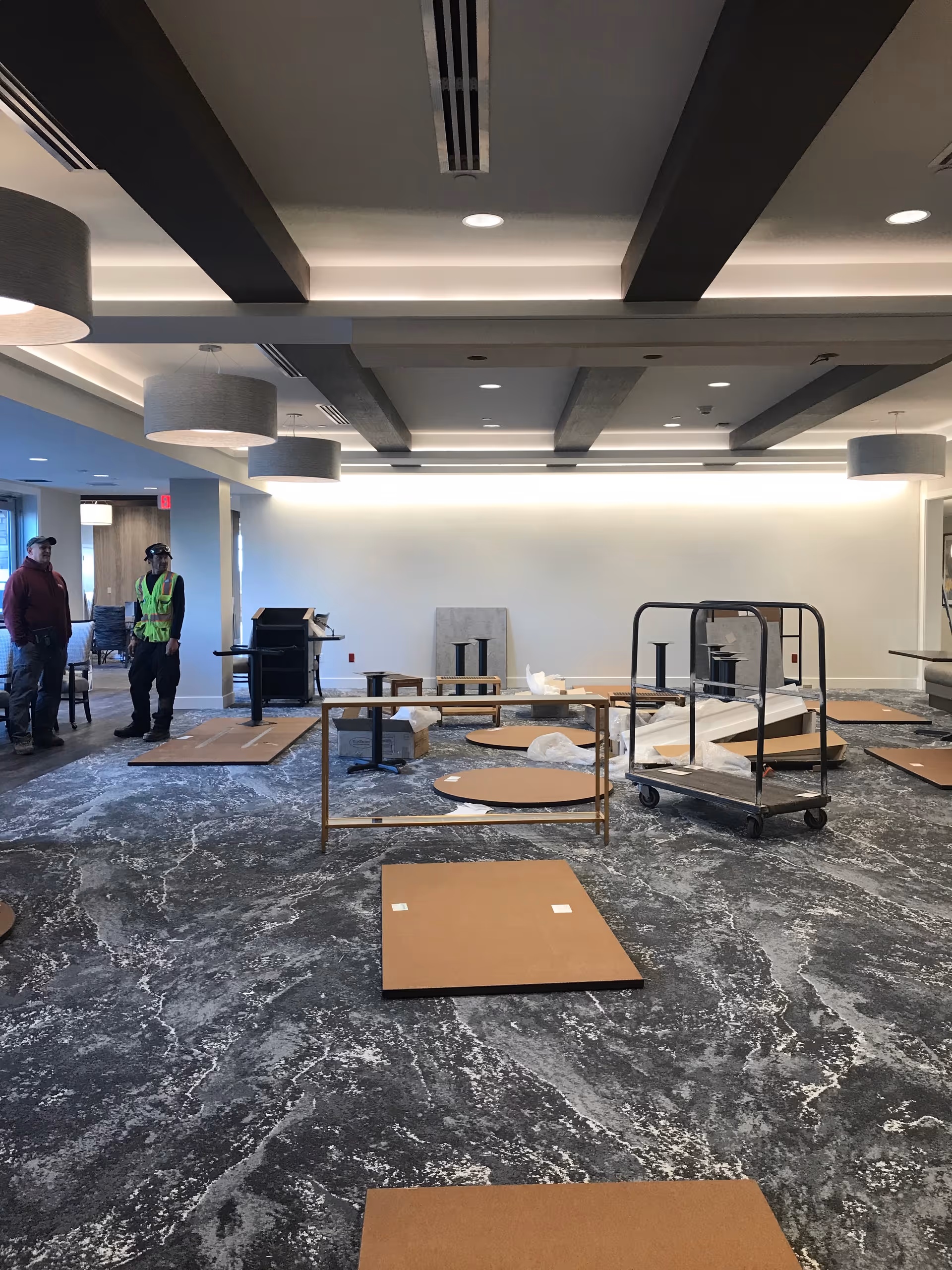 Spacious communal interior room being furnished with disassembled tabletops, carts and two workers standing at the left.