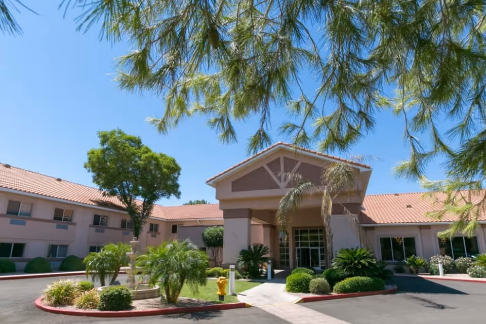 Front entrance of a senior living community building with a covered porte-cochère, palm trees, and a small fountain in a landscaped driveway.