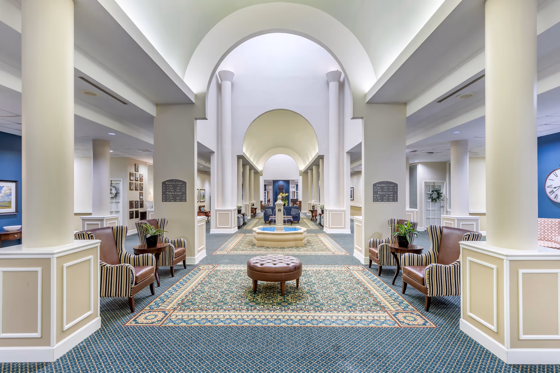 Spacious and elegant interior hallway of a senior living facility with high arched ceilings, white columns, patterned carpet, and seating areas featuring leather and striped armchairs with small tables and plants. A decorative fountain is centered in the distance.