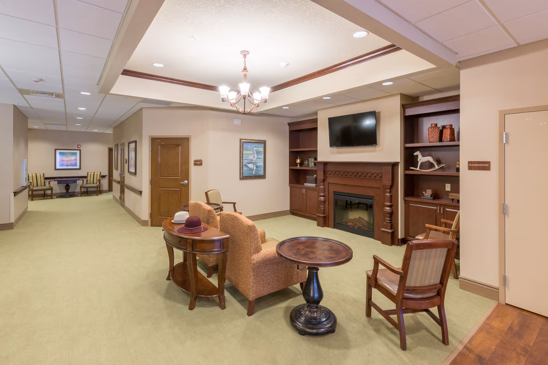 Cozy common lounge area with armchairs, small tables, a fireplace, built-in shelving and a wall-mounted TV in a senior living facility.