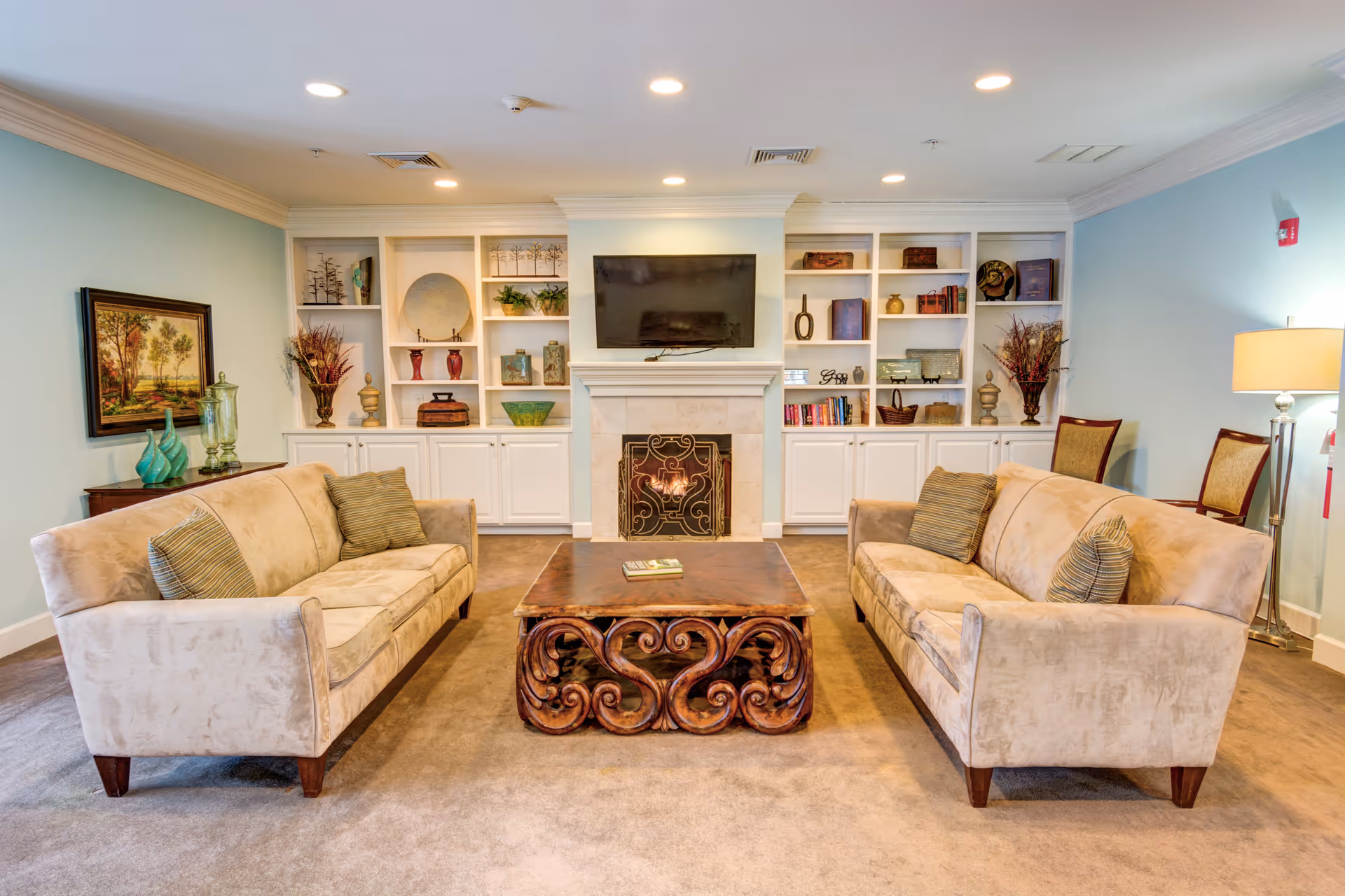 Bright common living room with two sofas facing a carved coffee table, built-in shelves and a wall-mounted TV above a fireplace.