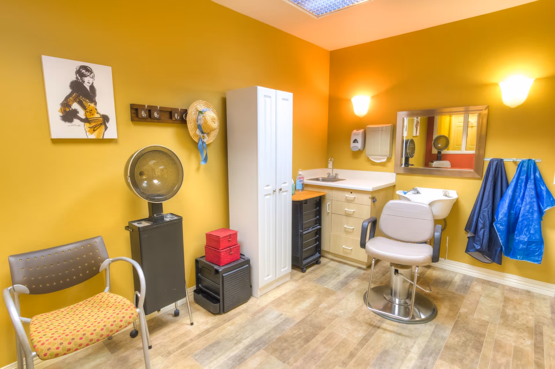 Interior of a small hair salon or beauty room with mustard yellow walls, a salon chair in front of a sink, a mirror, a hair dryer, a chair with a patterned cushion, a white storage cabinet, and hairdressing capes hanging on hooks.