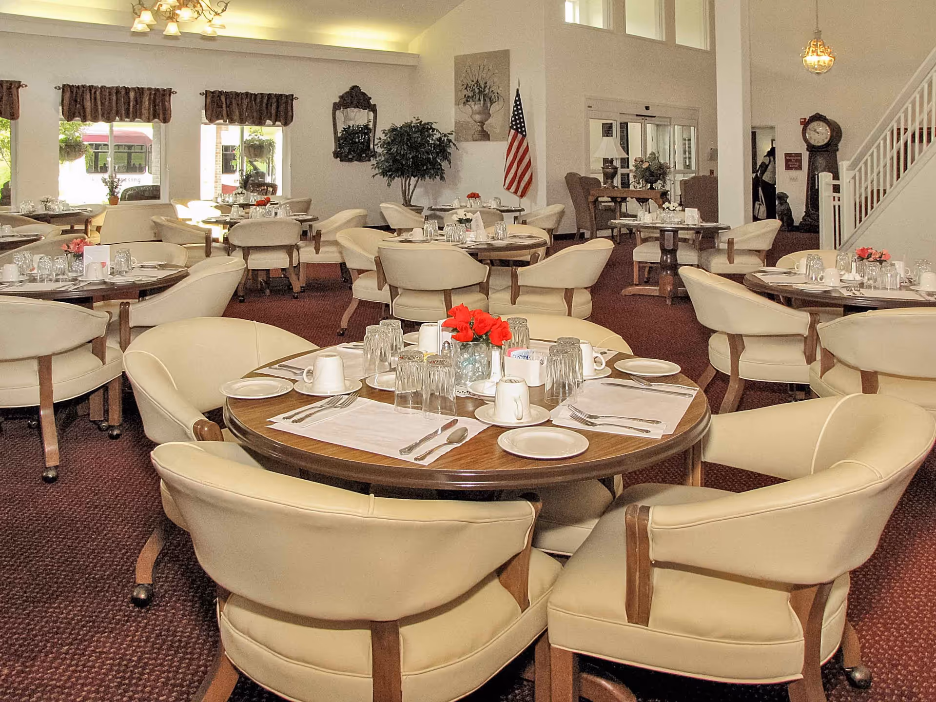 A dining room in a senior living facility with multiple round tables set with placemats, plates, cups, glasses, and silverware. Each table has a small flower arrangement in the center. The room has beige cushioned chairs, carpeted floor, large windows with valances, and an American flag near the entrance. There is a staircase and a grandfather clock in the background.