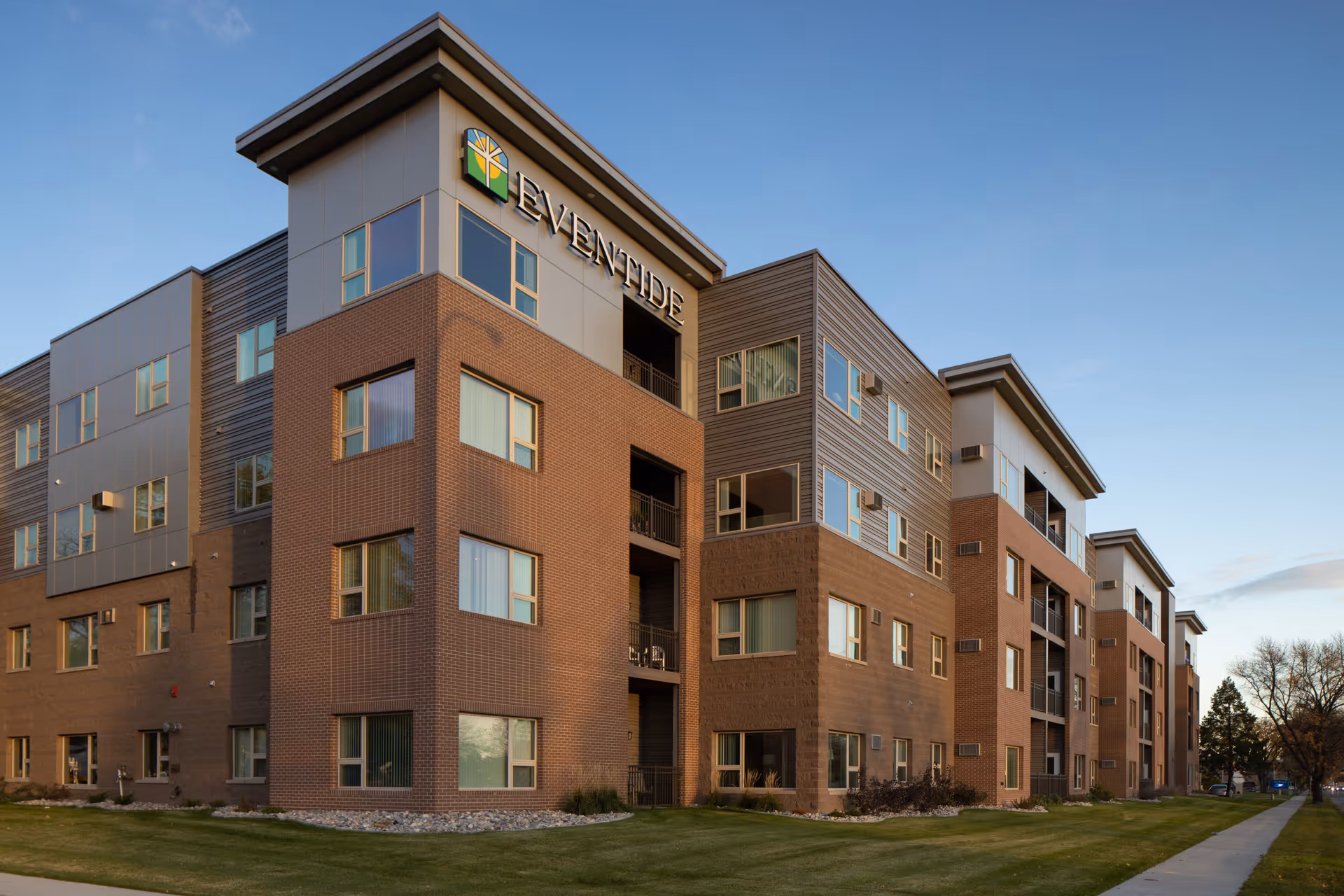 Exterior view of a modern multi-story senior living facility building named Eventide, with a combination of brick and metal siding, multiple windows, balconies, and a well-maintained lawn and sidewalk in front under a clear blue sky.