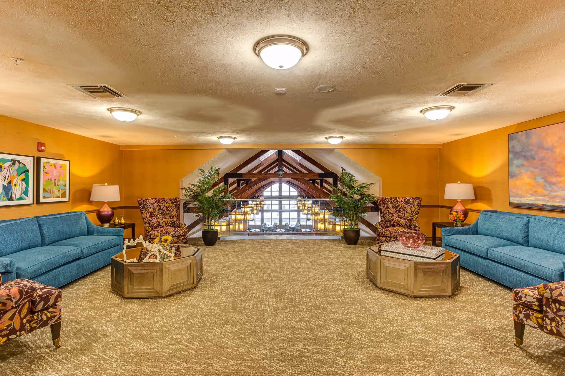 Spacious and well-lit lounge area with two blue sofas, two floral armchairs, two wooden coffee tables, potted plants, and colorful artwork on the walls. The room features a large window with wooden beams and hanging lights visible through the opening.