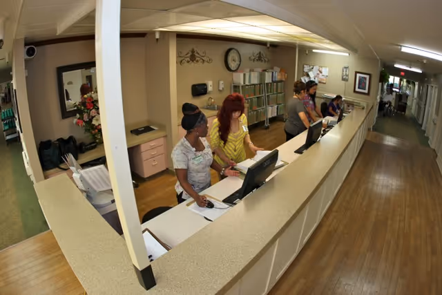 Reception area inside Upland Rehabilitation & Care Center with four staff members working behind a long counter. The area includes computer monitors, paperwork, a clock on the wall, and file organizers. A hallway extends to the right with additional rooms and a person in a wheelchair visible in the distance.