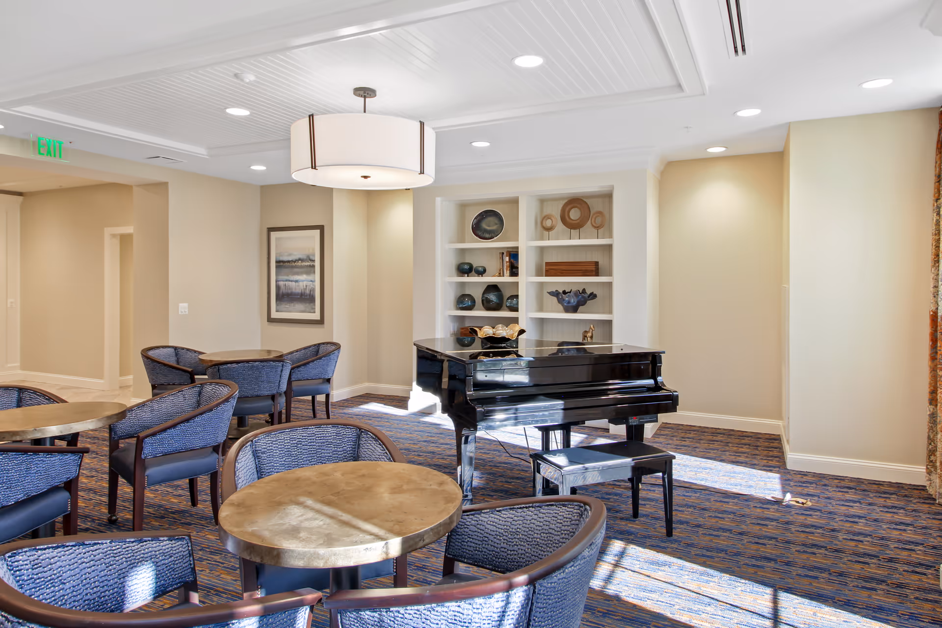 A bright senior living common area with round tables and blue cushioned chairs, a black grand piano with a matching bench, built-in shelves with decorative items, a framed painting on the wall, and a large ceiling light fixture.