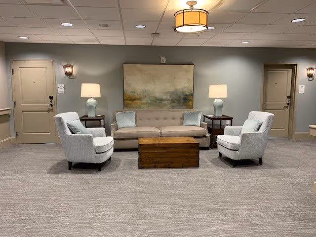 A cozy seating area in a senior living facility hallway with a beige sofa, two light gray armchairs, a wooden coffee table, two side tables with matching lamps, and a large abstract painting on the wall. There are two doors on either side of the seating area, wall sconces, and a ceiling light fixture.