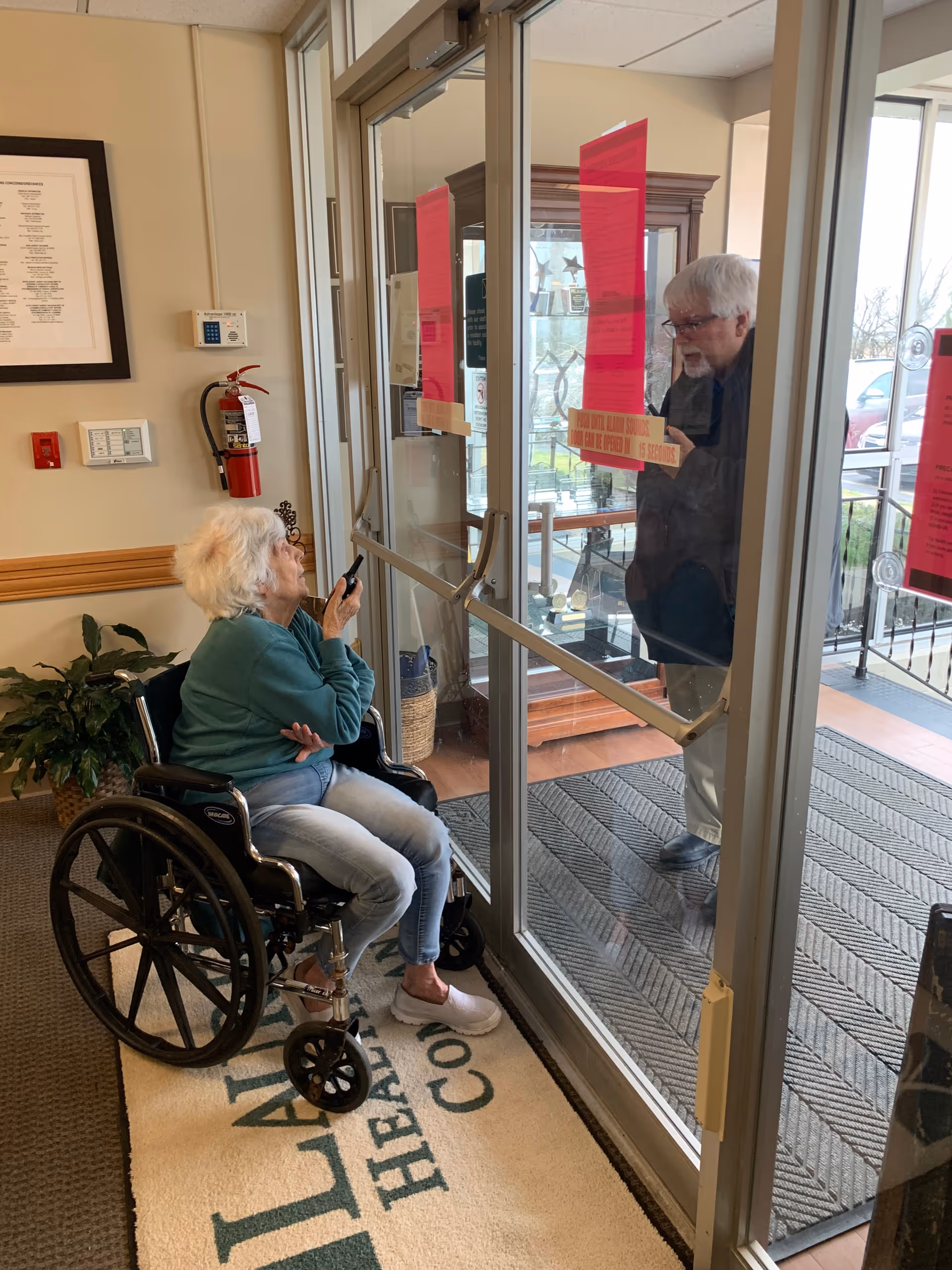 An elderly woman in a wheelchair inside a building is holding a phone and looking through a glass door at an elderly man standing outside. The interior has a fire extinguisher mounted on the wall, a framed document, a plant, and a mat on the floor with the text 'Laurels Health Care'.
