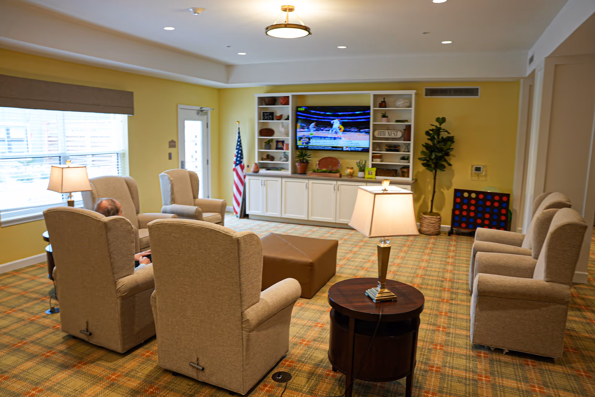 Bright communal living room with upholstered armchairs arranged around a central ottoman, a wall-mounted TV, and built-in shelving.