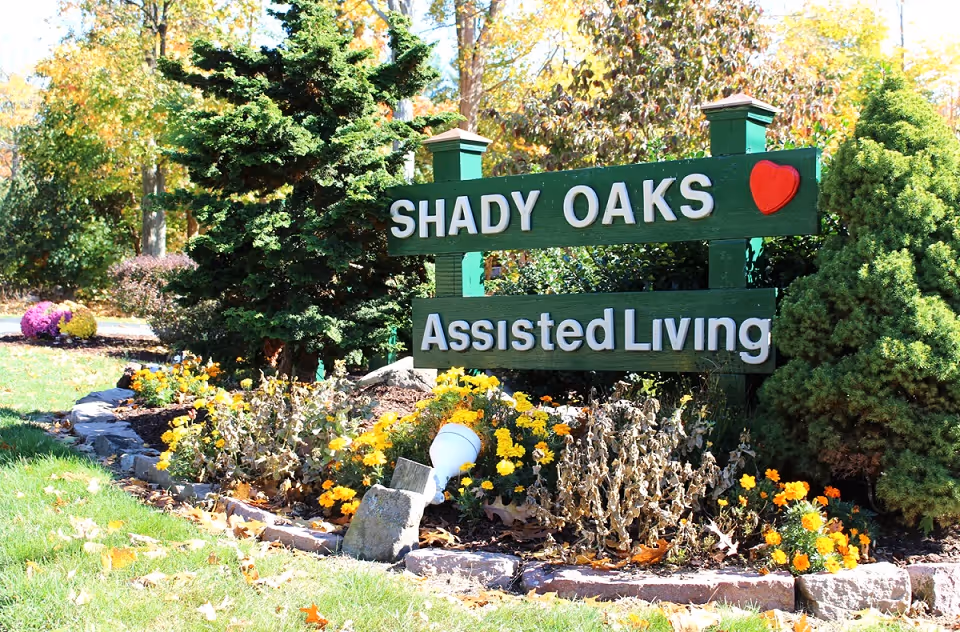 A green wooden sign with white letters reading 'SHADY OAKS Assisted Living' with a red heart symbol on the top right corner, surrounded by a landscaped garden with yellow and orange flowers, shrubs, and trees.