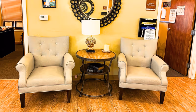 Two beige upholstered armchairs with button tufting are positioned on either side of a round wooden side table. The table holds a decorative lamp and a candle. Behind the table is a circular mirror with a decorative frame mounted on a yellow wall. To the left and right of the chairs are doorways leading to other rooms, one with a sign labeled 'Human Resources'.