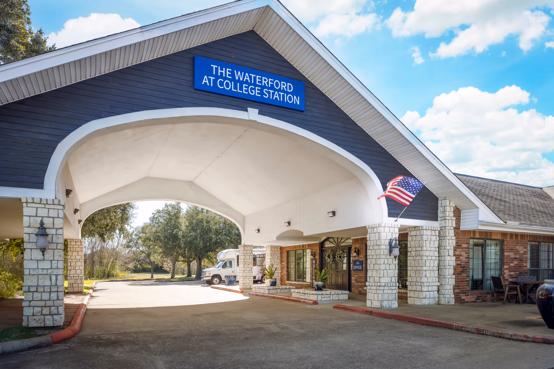 Front entrance canopy of The Waterford at College Station with stone pillars, driveway, and an American flag.