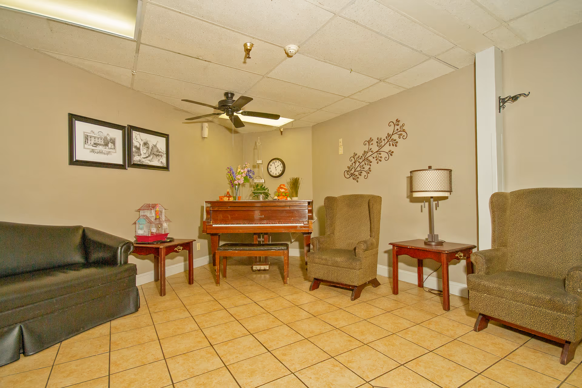 A cozy interior room with beige tiled flooring and light beige walls. The room features a black leather sofa on the left, two brown upholstered armchairs on the right, and a wooden piano with a bench in the center back. There are two framed black and white pictures on the left wall, a decorative wall decal on the right wall, a table lamp on a wooden side table, and a ceiling fan with lights above. A clock and some decorative items are placed on the piano.