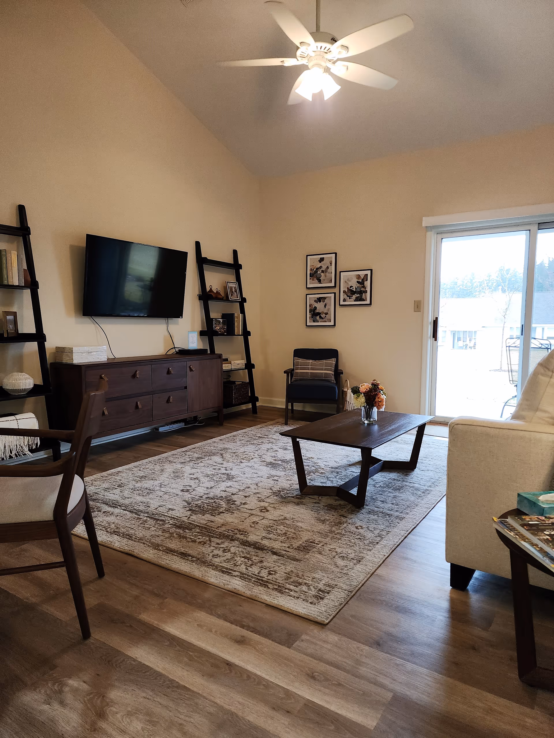 A cozy living room with a ceiling fan, a wall-mounted flat-screen TV, two black ladder-style shelves with books and decor, a dark wooden media console, a patterned area rug, a wooden coffee table with a small flower arrangement, a beige sofa, a wooden armchair with light cushions, and a sliding glass door leading outside.