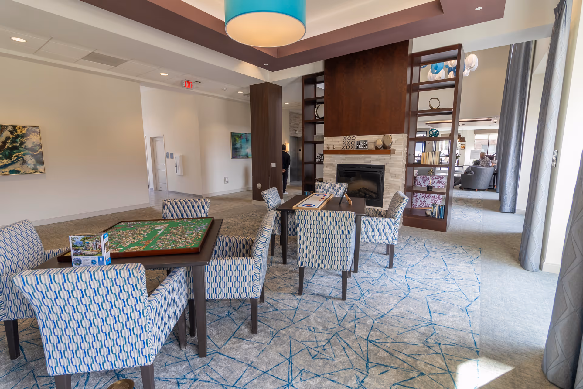 A bright and spacious common area in a senior living facility featuring two tables with patterned chairs around them. One table has a puzzle in progress and a puzzle box, while the other has a shuffleboard game. The room has a modern fireplace with decorative shelving on either side, abstract artwork on the walls, and large windows with curtains allowing natural light to fill the space. A person is visible in the background near the seating area.
