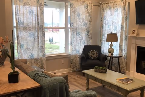 A cozy living room with a large window covered by floral curtains, a dark armchair with a decorative pillow, a round side table with a lamp, a light green coffee table with a small plant and magazines, and a sofa with a green throw blanket. A fireplace and a mounted TV are visible on the right side.