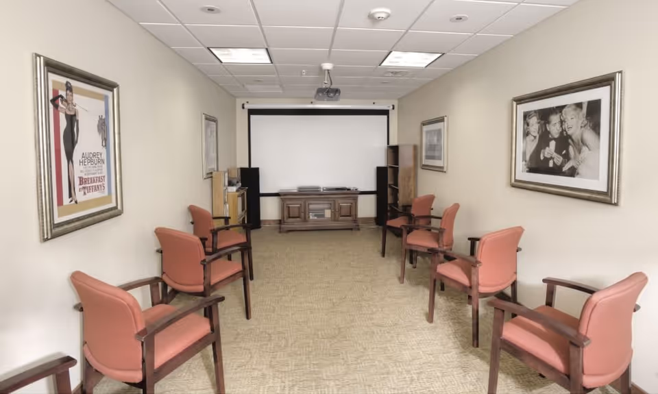 A small screening room with eight pink cushioned chairs arranged in two rows facing a large white projection screen. The room has beige walls adorned with framed black and white movie posters, including one of Audrey Hepburn in Breakfast at Tiffany's. There is a wooden cabinet under the screen and bookshelves on either side.