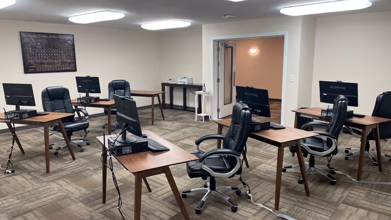 A computer lab with six wooden desks arranged in two rows, each desk equipped with a computer monitor, keyboard, and mouse. Each desk has a black leather office chair. The room has beige walls, carpeted floor with a geometric pattern, and bright overhead lighting. A framed periodic table of elements hangs on one wall, and there is a small table with a printer and hand sanitizer near an open door.