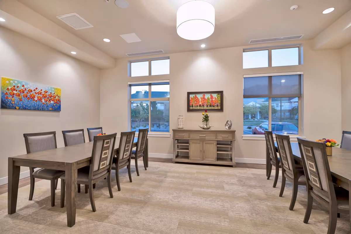 A bright dining room with two long wooden tables surrounded by chairs. The room has beige walls, carpeted floor, and large windows letting in natural light. There are colorful paintings on the walls and a sideboard with decorative items including a vase with a single red rose.