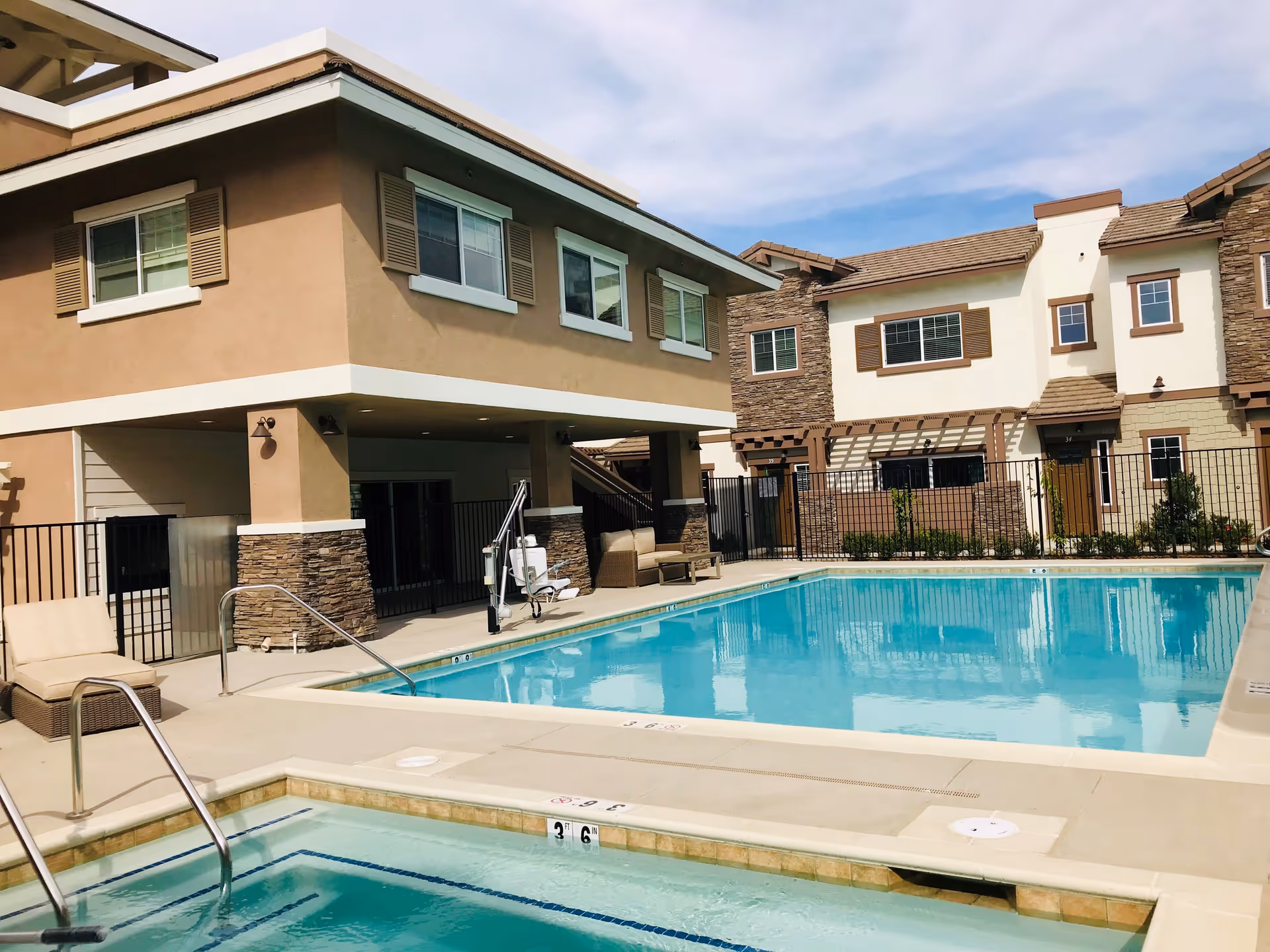 Outdoor swimming pool and hot tub area at a residential community with beige and brown two-story buildings in the background under a partly cloudy sky.